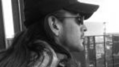 Wearing Sunglasses and Usual Black Ball Cap With Long Hait Looking Over Los Angeles From A Tall Building.
